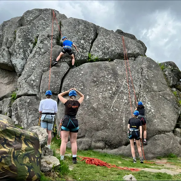 Vier mensen met helmen en klimgordels zijn aan het rotsklimmen en elkaar aan het zekeren op een groot, rotsachtig terrein in de open lucht onder een bewolkte hemel; op de grond liggen touwen en klimuitrusting.